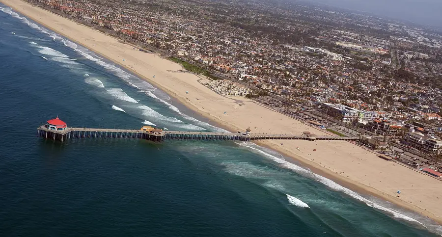 Aerial view of the Huntington Beach pier reaching out over the surf, with the wide beach and the downtown blocks along Main Street behind it.